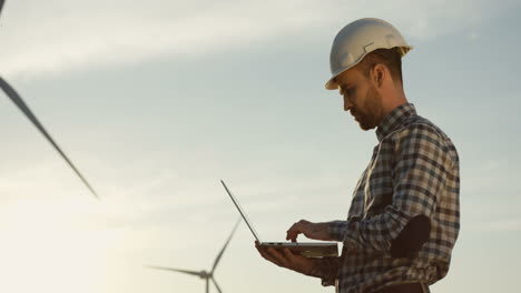 Side-view-of-Caucasian-man-engineer-wearing-a-helmet-using-laptop-while-checking-operation-of-the-windmill-tourbines