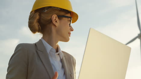 Lower-view-of-Caucasian-woman-engineer-wearing-a-helmet-using-laptop-while-checking-operation-of-the-windmill-tourbines