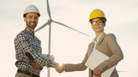 Lower-view-of-Caucasian-woman-and-man-engineers-wearing-a-helmet-shaking-hands-and-looking-at-camera-at-wind-station-of-renewable-energy