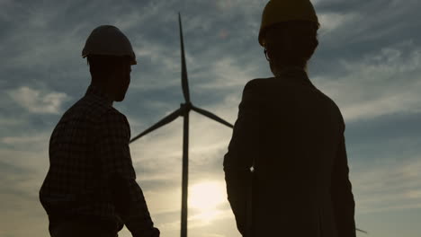 Silhouettes-of-a-caucasian-woman-and-man-engineers-wearing-a-helmet-shaking-hands-and-looking-at-camera-at-wind-station-of-renewable-energy