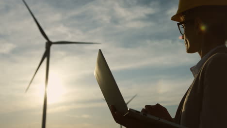 Silhouette-of-caucasian-woman-engineer-wearing-a-helmet-and-using-laptop-at-wind-station-of-renewable-energy