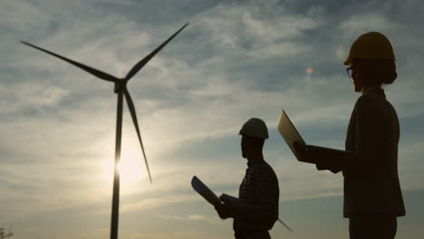 Silhouettes-of-a-caucasian-woman-and-man-engineers-wearing-a-helmet-and-reading-some-documents-or-blueprints-at-wind-station-of-renewable-energy