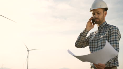 Caucasian-man-engineer-wearing-a-helmet-talking-on-the-phone-and-watching-some-blueprints-at-wind-station-of-renewable-energy