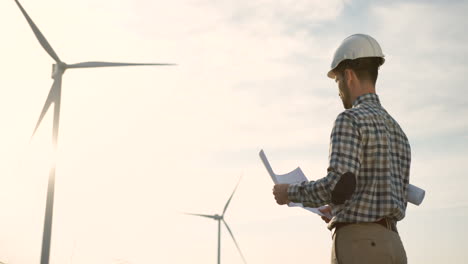Rear-view-of-caucasian-man-engineer-wearing-a-helmet-and-watching-some-blueprints-at-wind-station-of-renewable-energy