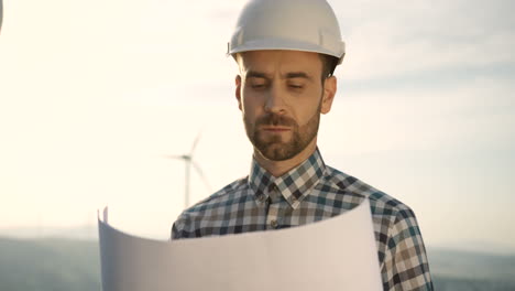 Close-up-view-of-caucasian-man-engineer-wearing-a-helmet-and-watching-some-blueprints-at-wind-station-of-renewable-energy