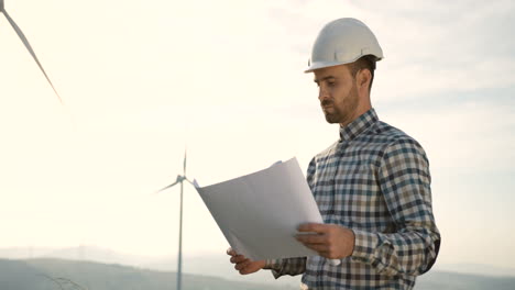 Caucasian-man-engineer-wearing-a-helmet-and-watching-some-blueprints-at-wind-station-of-renewable-energy