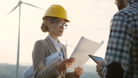 Caucasian-man-and-woman-engineers-wearing-a-helmet-watching-some-blueprints-and-using-tablet-while-talking-at-wind-station-of-renewable-energy
