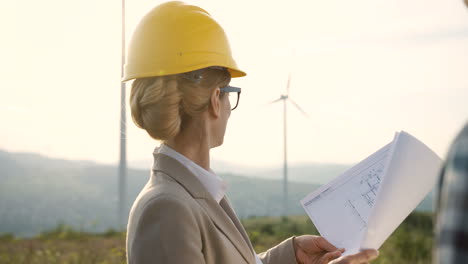 Caucasian-woman-engineer-wearing-a-helmet-watching-some-blueprints-and-talking-with-her-coworker-at-wind-station-of-renewable-energy