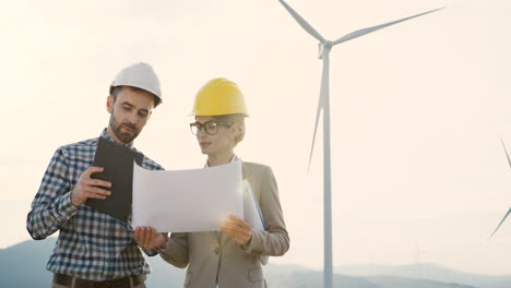 Caucasian-man-and-woman-engineers-wearing-a-helmet-watching-some-blueprints-and-using-tablet-while-talking-at-wind-station-of-renewable-energy