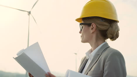 Close-up-view-of-caucasian-woman-engineer-wearing-a-helmet-watching-some-blueprints-at-wind-station-of-renewable-energy