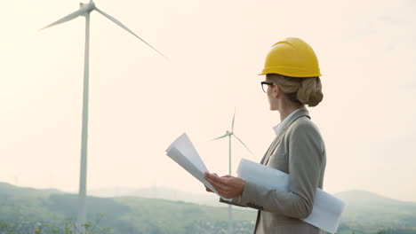 Caucasian-woman-engineer-wearing-a-helmet-watching-some-blueprints-at-wind-station-of-renewable-energy