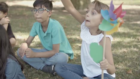 Cute-Caucasian-girl-holding-paper-fan,-catching-football-ball-and-throwing-it-away-while-sitting--with-friends-on-grass-in-the-park
