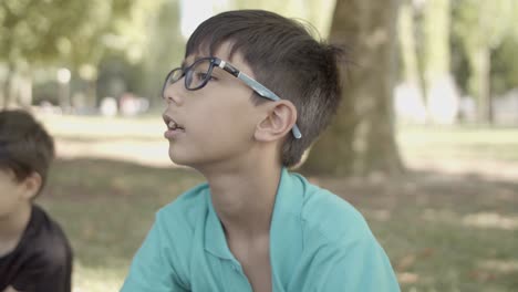 Portrait-of-Latin-boy-with-eyeglasses-sitting-on-grass-in-the-park,-listening-someone-and-talking
