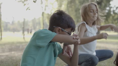 Cute-boy-in-eyeglasses-rubbing-nose-with-elbow-and-talking-with-girl-while-they-sitting-on-grass-in-the-park