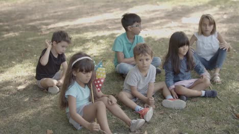 Niños-Multiétnicos-Sentados-En-El-Césped-Del-Parque,-Escuchando-A-Alguien-Y-Hablando