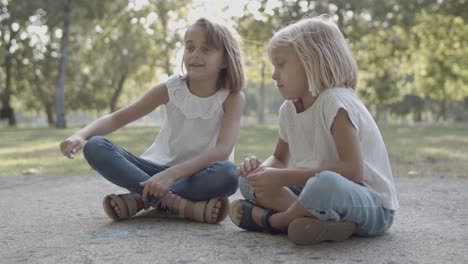 Two-Caucasian-girls-sitting-cross-legged-and-drawing-with-chalks-on-asphalt