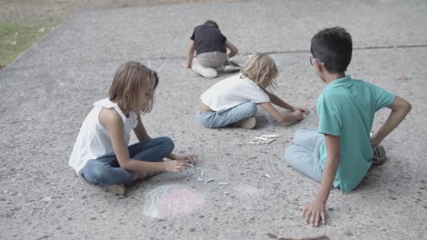 Multiethnic-children-sitting-on-asphalt-and-drawing-with-chalks