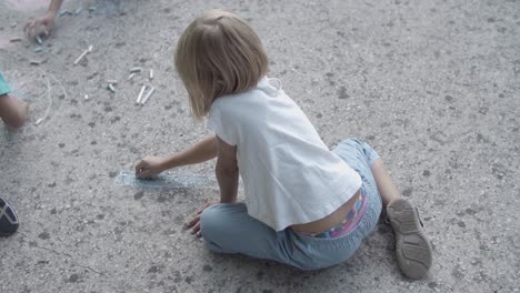 Back-view-of-blonde-girl-sitting-on-asphalt-and-drawing-with-chalks