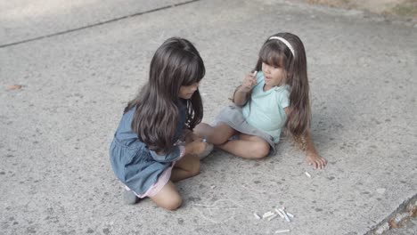Two-Latin-little-girls-sitting-on-asphalt,-drawing-with-chalks-and-talking