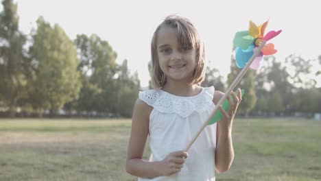 Portrait-of-a-smiling-Caucasian-girl-holding-paper-fan,-jumping-and-looking-at-the-camera-in-the-park