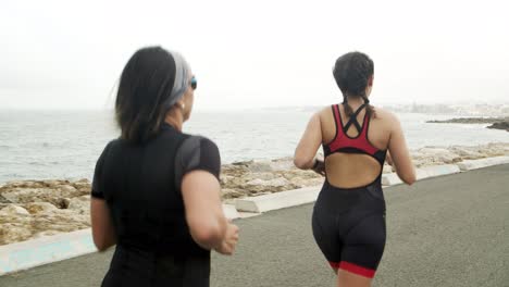 Vista-Trasera-De-Triatletas-Femeninas-Entrenando-Para-Maratón-En-Una-Carretera-Cerca-Del-Mar