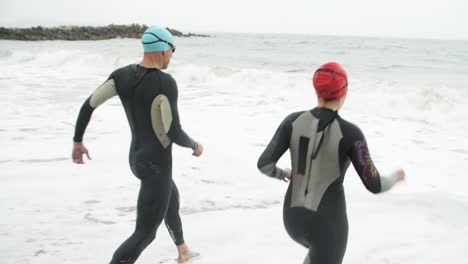 Athletic-man-and-woman-running-on-beach-with-wetsuits
