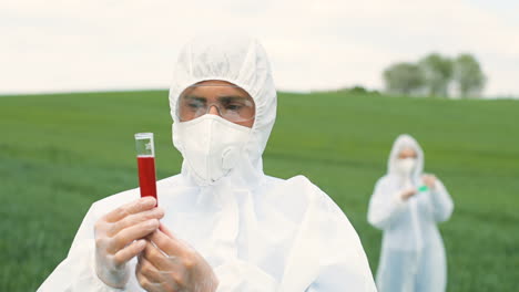 Close-up-view-of-caucasian-researcher-man-in-protective-suit-holding-test-tube-while-doing-pest-control-in-the-green-field