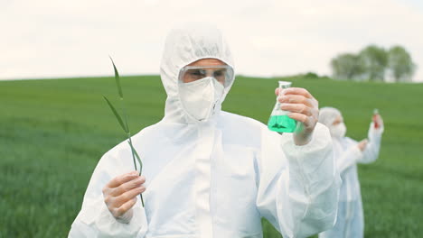 Close-up-view-of-caucasian-researcher-man-in-protective-suit-holding-test-tube-and-tree-branch-while-doing-pest-control-in-the-green-field