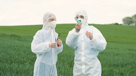 Caucasian-researchers-in-protective-suit-holding-test-tubes-and-tree-branch-while-doing-pest-control-in-the-green-field