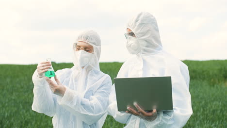 Caucasian-researchers-in-protective-suit-holding-a-test-tube-and-using-laptop-while-doing-pest-control-in-the-green-field