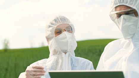 Close-up-view-of-caucasian-researchers-in-protective-suit-holding-a-test-tube-and-using-laptop-while-doing-pest-control-in-the-green-field