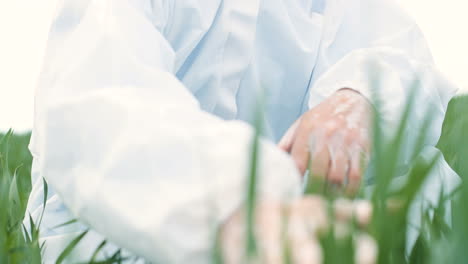 Close-up-view-of-Caucasian-researcher-man-in-protective-suit-and-goggles-sitting-on-green-field-and-touching-organic-herb-of-wheat