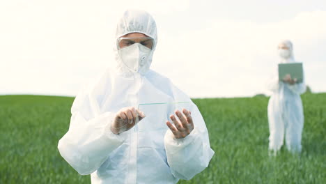 Caucasian-researcher-man-in-protective-suit-and-goggles-tapping-on-glass-in-the-green-field