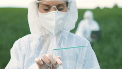 Rear-view-of-researcher-woman-in-protective-suit-and-goggles-tapping-on-glass-in-the-green-field