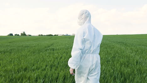 Rear-view-of-caucasian-researcher-man-in-white-protective-suit-and-goggles-walking-in-the-green-field-while-doing-pest-control