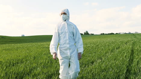 Caucasian-researcher-man-in-white-protective-suit-and-goggles-walking-in-the-green-field-while-doing-pest-control