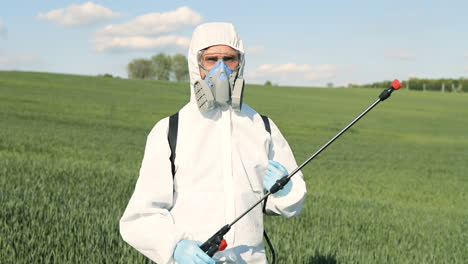 Close-up-view-of-caucasian-researcher-man-in-white-protective-suit-and-goggles-holding-pulverizator-in-the-green-field-while-looking-at-camera