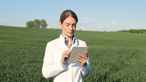 Caucasian-researcher-woman-in-white-coat-using-tablet-and-looking-at-camera-in-the-green-field