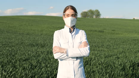 Caucasian-researcher-woman-in-white-coat-and-goggles-looking-at-camera-with-arms-crossed-in-the-green-field