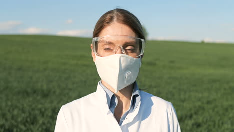 Close-up-view-of-caucasian-researcher-woman-in-white-coat-and-goggles-looking-at-camera-in-the-green-field