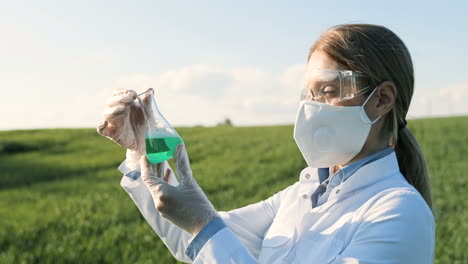 Close-up-view-of-caucasian-researcher-woman-in-white-coat-and-goggles-looking-at-test-tube-in-the-green-field