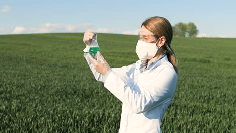 Caucasian-researcher-woman-in-white-coat-and-goggles-looking-at-test-tube-in-the-green-field