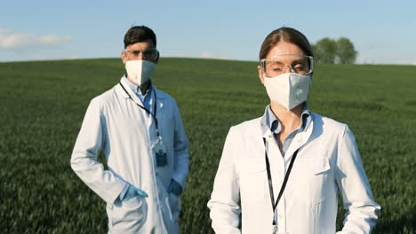 Caucasian-researchers-in-white-gown,-mask-and-googles-looking-at-camera-in-the-green-field