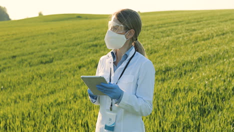 Caucasian-researcher-woman-in-white-coa,-mask-and-goggles-using-tablet-and-looking-around-in-the-green-field