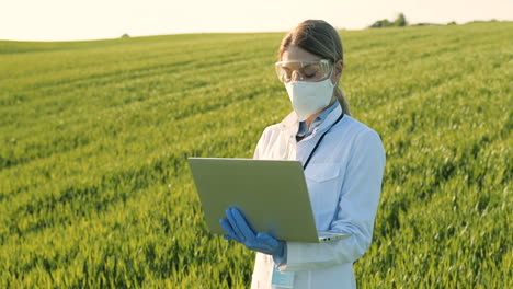 Caucasian-researcher-woman-in-white-coa,-mask-and-goggles-using-laptop-and-looking-around-in-the-green-field