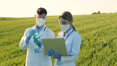 Kaukasische-Weibliche-Und-Männliche-Forscher,-Die-Reagenzgläser-Mit-Chemischen-Pestiziden-Halten-Und-Laptop-Im-Grünen-Feld-Verwenden