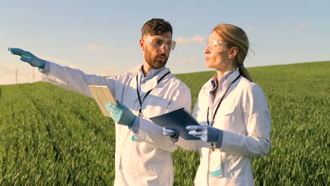 Caucasian-female-and-male-researchers-in-white-coat,-mask-and-googles-using-tablet-and-taking-notes-while-doing-pest-control-in-the-green-field