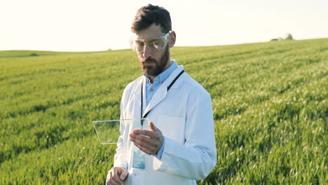 Caucasian-researcher-man-in-white-coat-and-goggles-tapping-on-glass-in-the-green-field