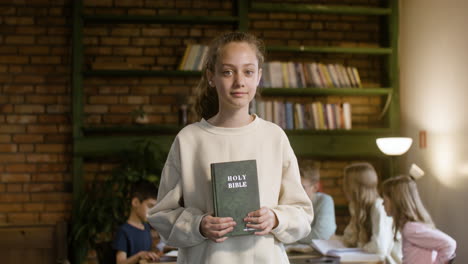 Young-student-holding-the-Bible-on-her-hands-while-her-classmates-study-behind-her