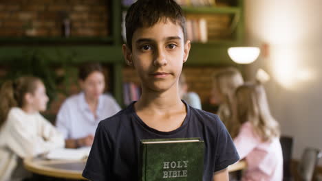 Brunette-young-student-showing-Bible-book-on-his-hands-at-class